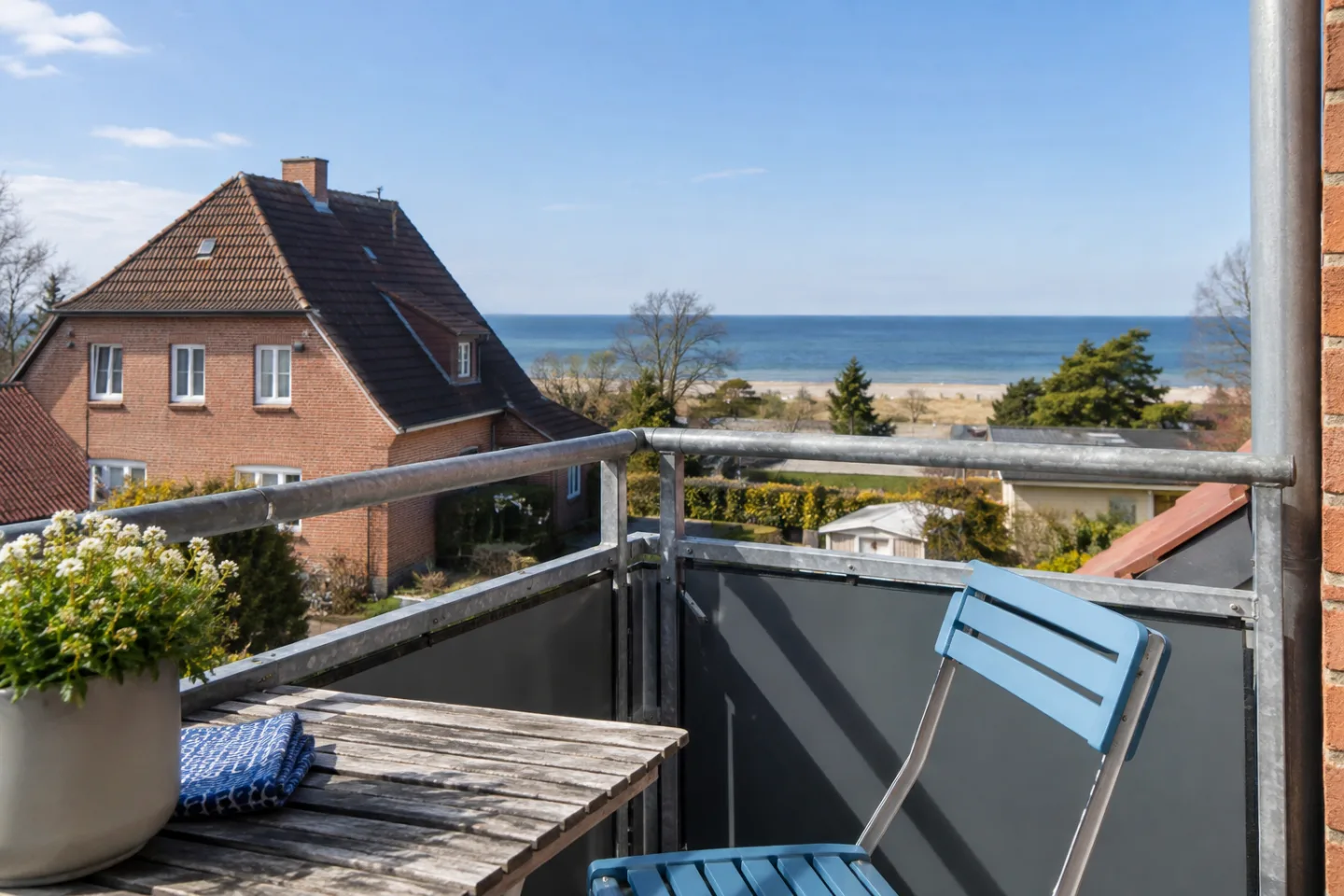 Balkon mit blauem Klappstuhl und Blick über ein Backsteinhaus auf das Meer.