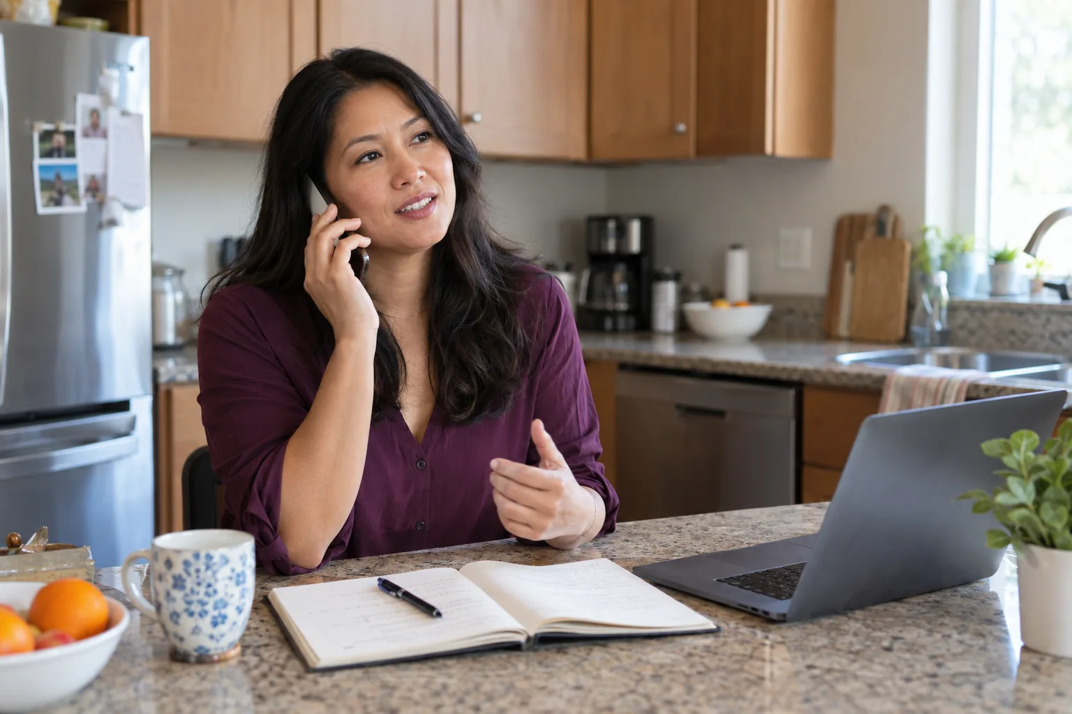 A woman speaks on the phone at a kitchen counter beside a laptop and notebook.